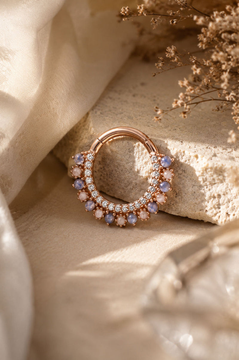 A rose gold hoop ring with rows of clear crystals and light blue oval gemstones, styled on a textured stone slab with draped cream fabric and dried flowers.